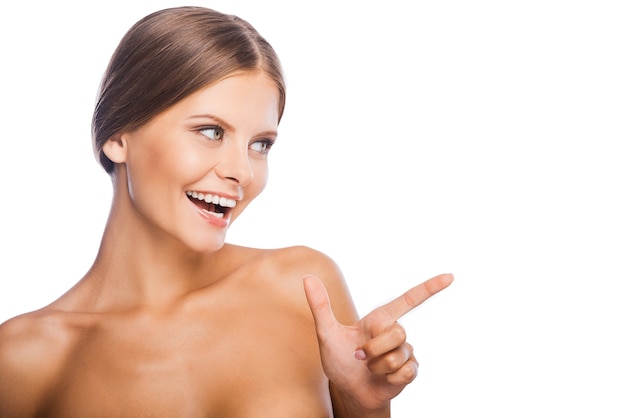 Schau dort! Fröhliche junge Frau mit nacktem Oberkörper, die weg zeigt und lächelt, während sie vor weißem Hintergrund steht
