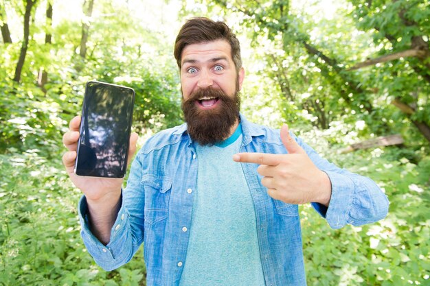 Schau hier. glücklicher bärtiger Mann, der mit dem Finger auf das Telefon zeigt. Reifer Hipster mit Bart. im Wald verloren. brutaler Mann mit Telefon. Hipster-Stil. Kerl im Wald verwendet GPS-Navigation. Sommercamping.