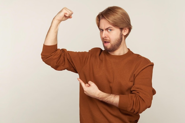 Schau, wie stark ich bin. Porträt eines selbstbewussten stolzen bärtigen Mannes im Sweatshirt, der Kraft in der Hand demonstriert und Bizeps zeigt