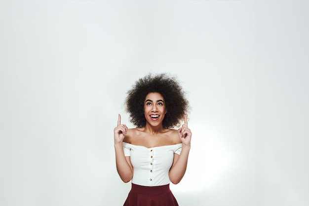 Schauen Sie dort hin Fröhliche und schöne afroamerikanische Frau mit lockigem Haar, die mit den Fingern nach oben zeigt, während sie vor grauem Hintergrund steht. Schönheit der Frauen. Studioportrait