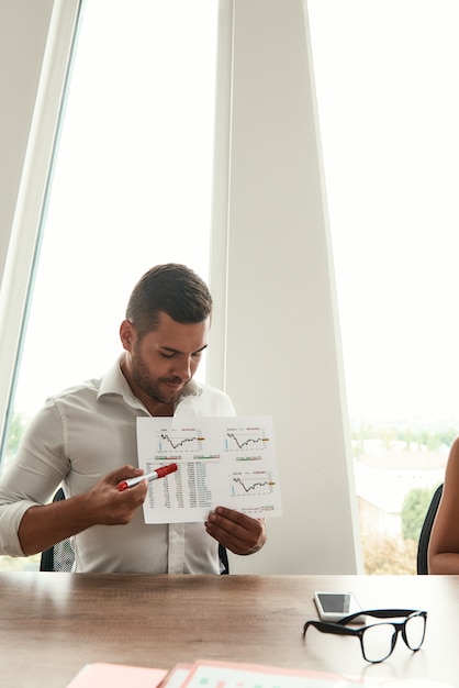 Schauen Sie hier, junger Mann in formeller Kleidung, der Finanzberichte zeigt, während er im Büro sitzt