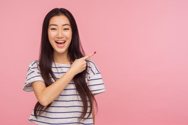 Schauen Sie sich die Werbung an, Porträt eines charmanten asiatischen Mädchens in gestreiftem T-Shirt, das zur Seite zeigt und lächelt, Kopierraum zeigt, leerer Platz für Werbung. Indoor-Studio-Shooting isoliert auf rosa Hintergrund