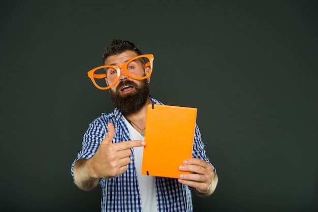 Schauen Sie sich dieses Buch an. Bärtiger Mann in Partybrille, der auf Lehrbuch zeigt. Studieren Sie Nerd, der Buch hält. Buch-Nerd mit schicker Brille. Männlicher Student der Universität mit Vorlesungsnotizen.