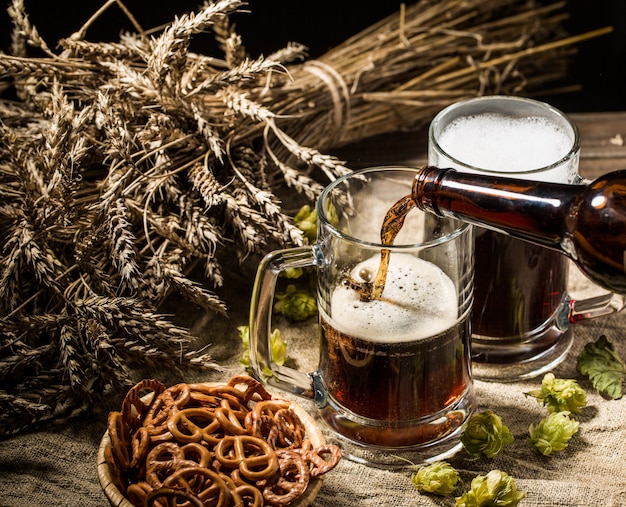 Schaumiges Bier aus der Flasche goss in einen Becher, der mit vollem Becher Bier mit Weizen und Hopfen, Korb Brezeln stand