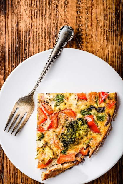 Scheibe Gemüsefrittata mit Brokkoli, rotem Paprika und roten Zwiebeln auf weißem Teller. Sicht von oben