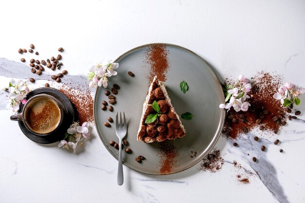 Scheibe hausgemachtes glutenfreies Tiramisu traditionelles italienisches Dessert mit Kakaopulver bestreut, blühender Apfelbaum, Kaffee, Minzblätter, Kaffeebohnen über weißer Marmoroberfläche. Ansicht von oben, flach