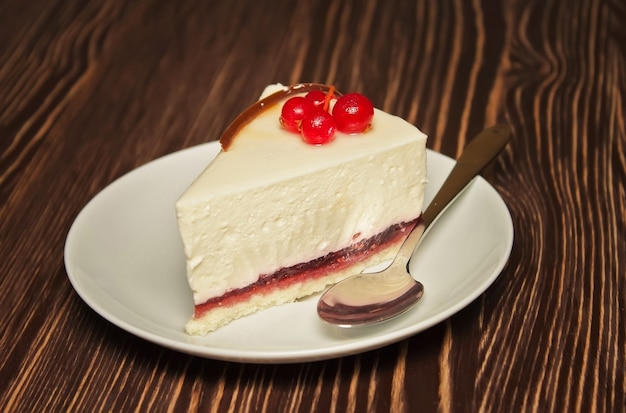 Scheibe Käsekuchen mit frischer roter Johannisbeere auf weißem Teller mit einem Teelöffel auf braunem hölzernem Hintergrund. Nahaufnahme