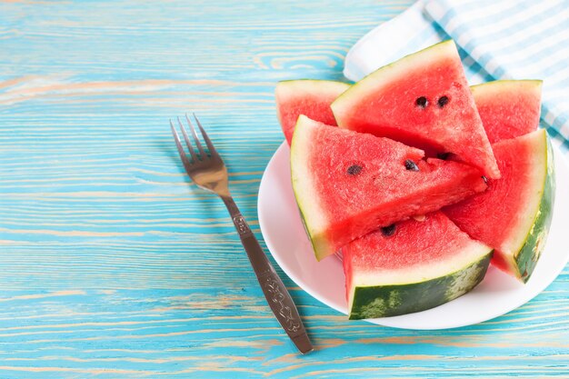 Scheiben der Wassermelone auf dem Teller auf blauem hölzernem Hintergrund mit Gabel und kariertem Handtuch.