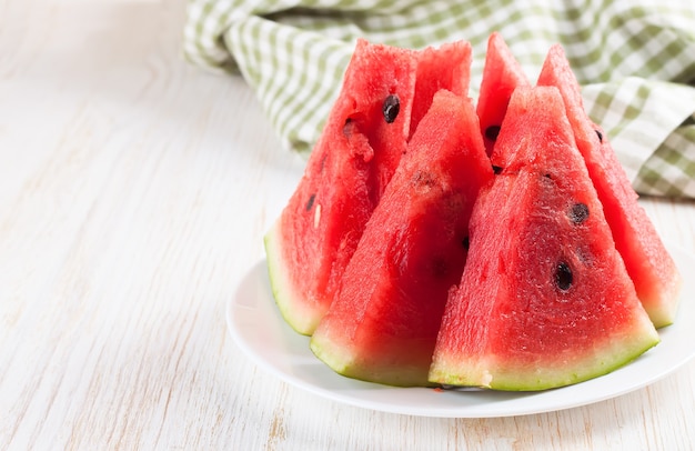 Scheiben der Wassermelone auf dem Teller auf weißem hölzernem Hintergrund mit Gabel und grünem kariertem Handtuch.