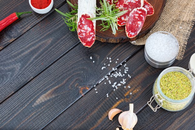 Scheiben gehackte Wurst, Gewürze in Gläsern und verstreut, Pfeffer Chili und Tomaten auf einem dunklen rustikalen Holzhintergrund.