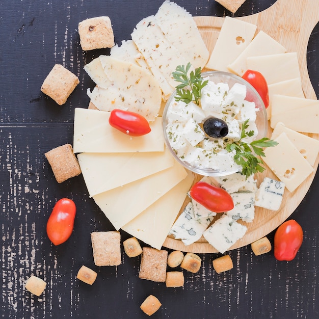 Foto scheiben käse auf dem hackenden brett umgeben mit tomaten und gebäck auf schwarzem strukturiertem hintergrund