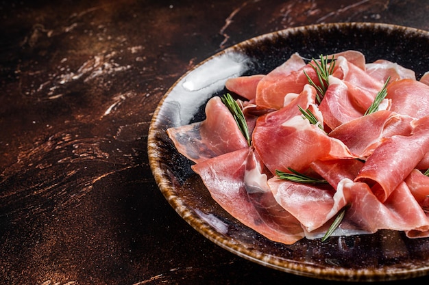 Scheiben Prosciutto Crudo Parma oder Jamon Serrano mit Rosmarin. Dunkler Hintergrund. Ansicht von oben. Platz kopieren.