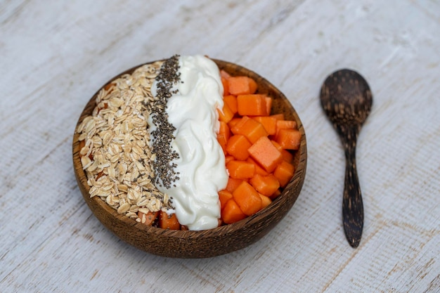 Scheiben süßer Papaya mit Haferflocken, Chiasamen und weißem Joghurt in Kokosnussschale auf weißem Holzhintergrund hautnah