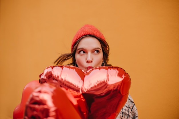 Foto schelmisches mädchen versteckt sich hinter rotem herzförmigem ballon nahaufnahmeporträt des jungen modells im hellen hut auf orangefarbenem hintergrund