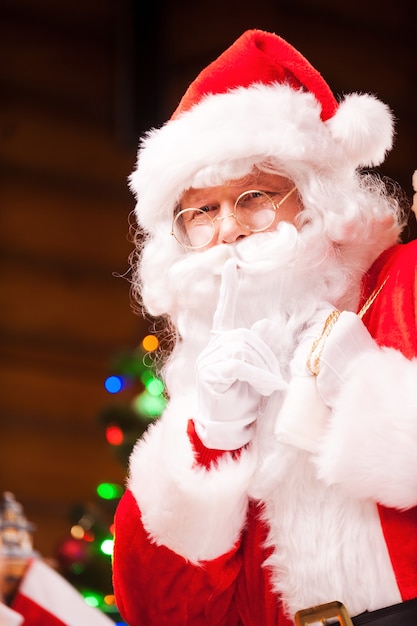 Schhh! Traditioneller Weihnachtsmann, der ein Stillezeichen gestikuliert, während er einen Sack mit Geschenken und einen Weihnachtsbaum im Hintergrund trägt