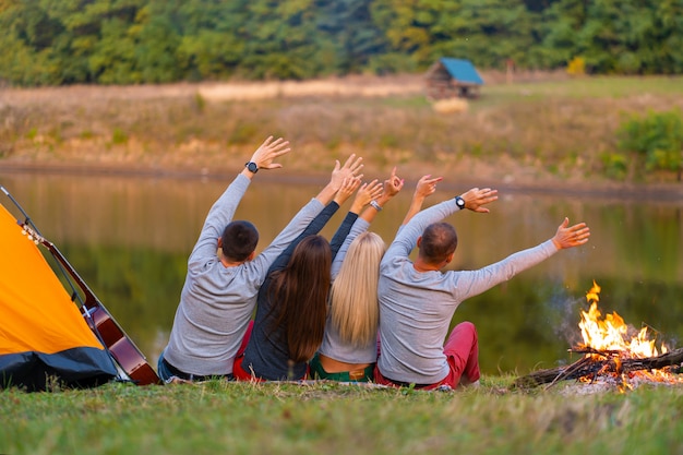 Schießen Sie von hinten. Eine Gruppe glücklicher Freunde, die am Flussufer kampieren, Händchen halten und Aussicht genießen. Urlaubsspaß