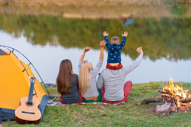 Schießen Sie von hinten. Eine Gruppe glücklicher Freunde mit Kind auf der Schulter kampierend am Flussufer, Tanzen halten Hände hoch und genießen Ansicht. Familienurlaub Spaß