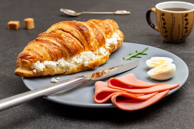 Schinken, Eier, Messer und Croissant mit Hüttenkäse auf grauem Teller. Tasse Kaffee. Ansicht von oben. Schwarzer Hintergrund.