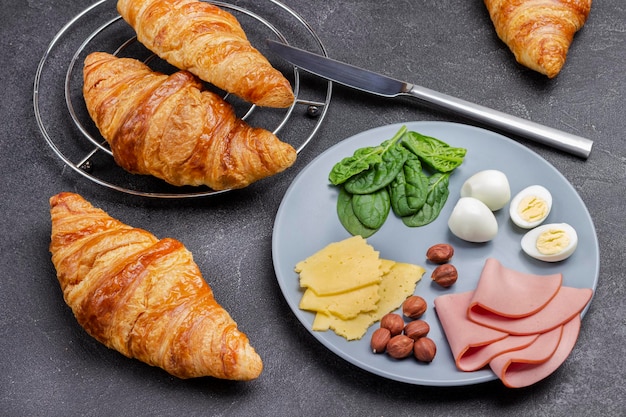 Schinken, Käse, Nüsse und Spinat auf grauem Teller. Frische Croissants und Messer. Ansicht von oben. Schwarzer Hintergrund.