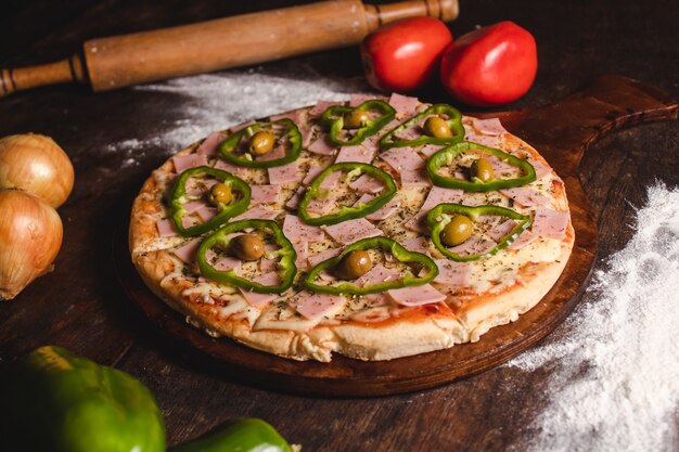 Schinken-Paprika-Pizza mit viel Käse und Gemüse rundum auf einem Holztisch.