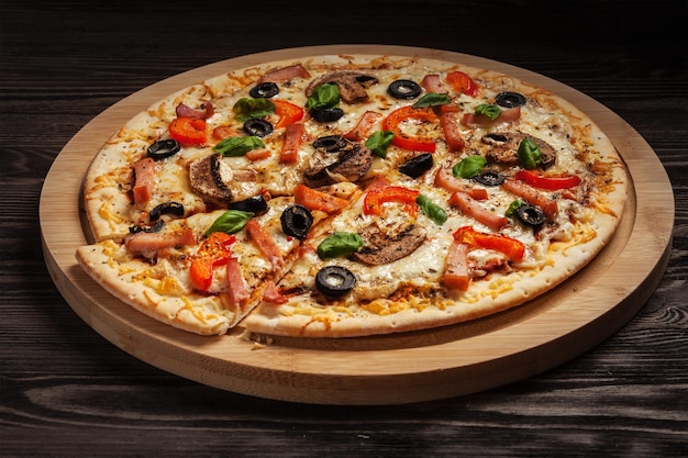 Schinken-Pizza mit Capsicum-Pilzen, Oliven und Basilikumblättern auf einem Holzbrett auf einem alten Tisch