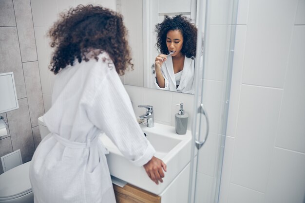 Schläfrige Dame mit einer Zahnbürste in der Hand, die an das Waschbecken im Badezimmer gelehnt ist