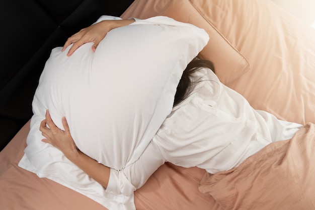 Schläfrige Frau versteckt sich unter dem Kissen hasst es, früh am Morgen aufzuwachen