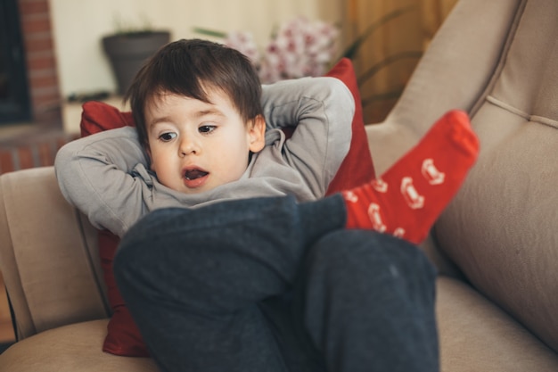 Schläfriger kaukasischer Junge mit roten Socken, die auf einem Sofa posierend liegen, bereit für ein Nickerchen