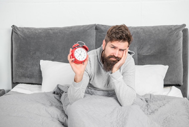 Schläfriger Mann mit Wecker, der morgens im Bett liegt