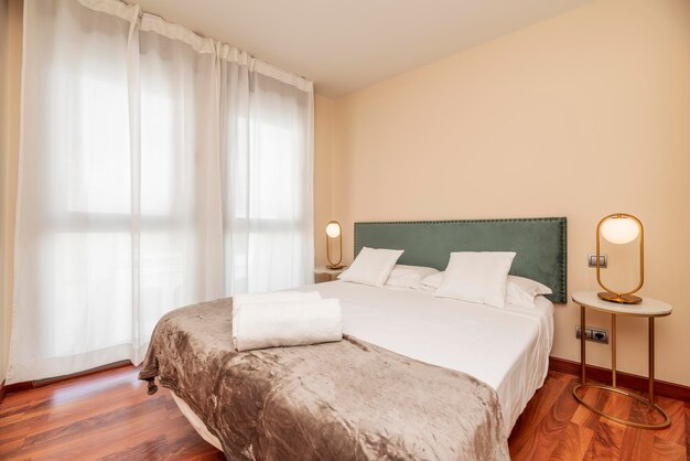 Schlafzimmer mit Doppelbett mit Kopfteil aus grünem Samtstoff und großen Fenstern mit weißen Vorhängen