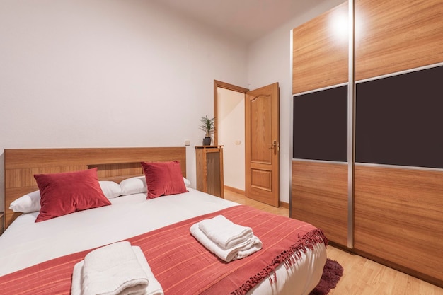 Schlafzimmer mit Kingsize-Bett, roten Kissen, passender Decke, weißer Bettwäsche und Schiebetürenschrank aus Holz