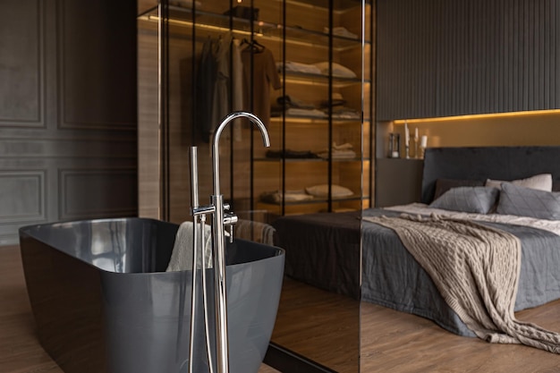 Schlafzimmer und freistehendes Bad hinter einer Glastrennwand in einem schicken, teuren Interieur eines Luxushauses mit dunklem, modernem Design mit Holzverkleidung und LED-Licht