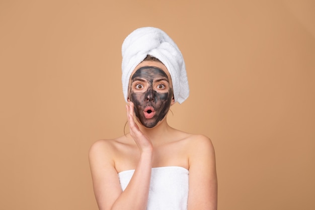 Schlamm-Gesichtsmaske, Gesichts-Ton-Maske Spa. Schöne Frau mit kosmetischer Schlammgesichtsbehandlung, Spa-Gesundheitskonzept. Hautpflege Schönheitsbehandlung. Handtuch auf dem Kopf. Medizinischer Mineralschlamm. Beige Hintergrund, isoliert.