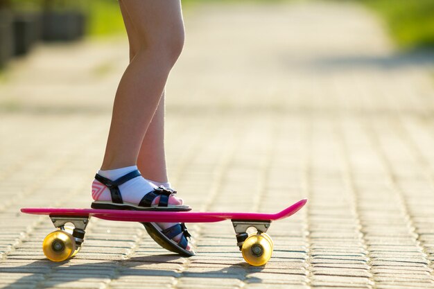 Schlanke Beine des Kindes in den weißen Socken und in den schwarzen Sandalen auf dem rosa Skateboard des Plastiks auf hellem sonnigem Sommer verschwommenem Kopienraumpflasterhintergrund. Outdoor-Aktivitäten und gesundes Lifestyle-Konzept.