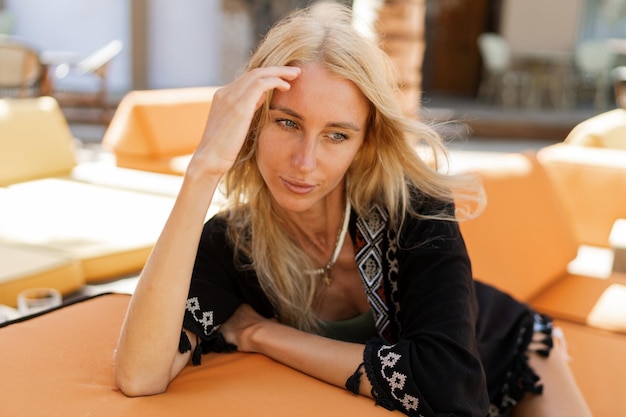 Schlanke, blonde Frau, die Urlaub genießt und im stilvollen Strandclub chillt. Tragen Sie ein stilvolles Boho-Sommeroutfit.