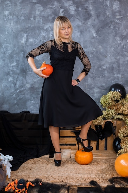 Schlanke blonde Frau im schwarzen kurzen Kleid und Absätzen mit Kürbissen unter dem Halloween-Dekor