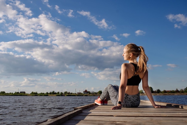 Schlanke Fitness-Frau sitzt und entspannt sich nach dem Training auf dem Pier bei Sonnenuntergang