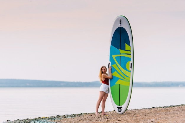 Schlanke Frau auf dem Strand mit Paddel-Sup-Board mit wunderschönen Sonnenuntergangs- oder Sonnenaufgangsfarben