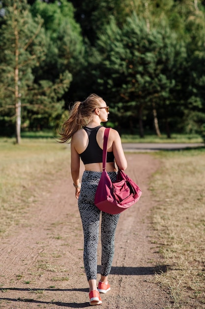 Schlanke Frau, die mit einer großen Sporttasche im Freien im Park spazieren geht