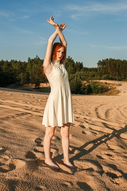 Schlanke Frau mit roten Haaren, die mit erhobenen Armen im Kleid am Sandstrand steht