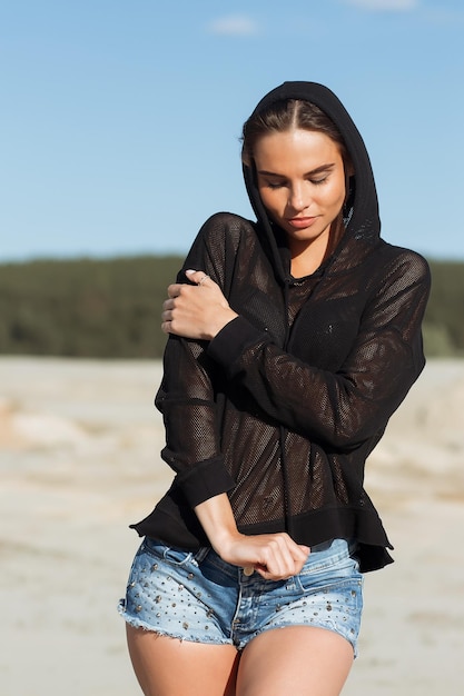 Schlanke, hinreißende Frau in Denim-Sommershorts und transparentem schwarzem Hoodie, die im Sonnenlicht an der Küste steht