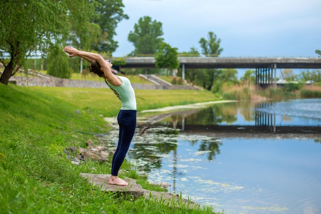 Schlanke junge Brünette Yogi führt keine komplizierten Yoga-Übungen auf dem grünen Rasen im Sommer