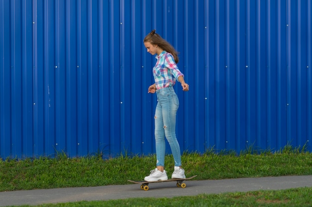 Schlanke junge Frau in Jeans aus dem Skateboardfahren entlang eines schmalen Pfades in der Stadt gegen eine bunte blaue Wand