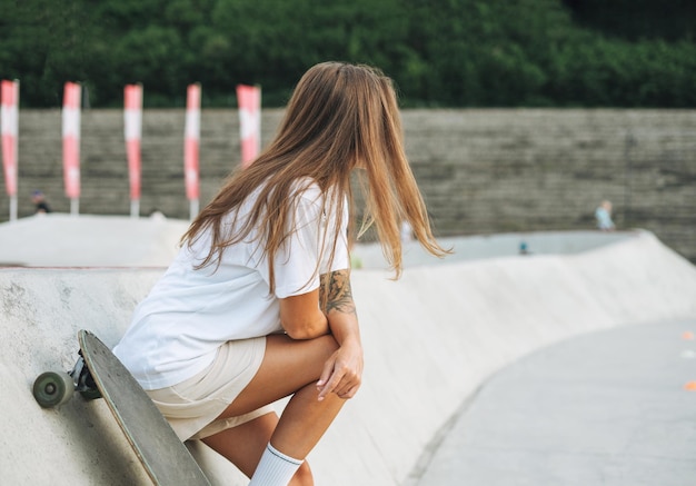 Schlanke junge Frau mit langen blonden Haaren in leichter Sportkleidung mit Longboard im Skatepark bei Sonnenuntergang