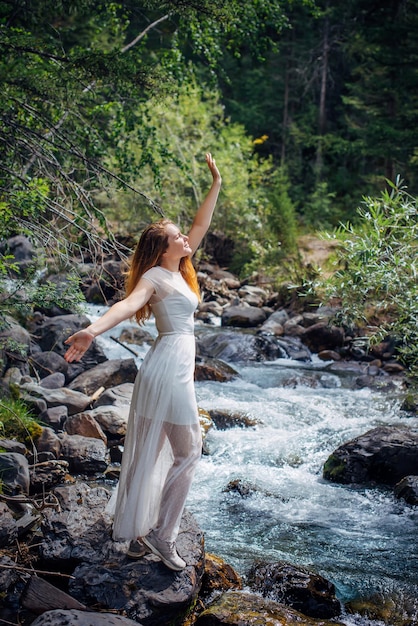 Schlanke langhaarige Brünette in weißem Kleid, die sich gegen kleine Gebirgsflüsse und grüne Bäume ausgibt Schöne junge Frau, die entlang des Waldstromufers vertikales Bild geht