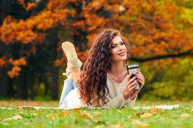 Schlanke schöne junge Frau ruht im Herbst im Park und liegt auf dem Rasen auf dem Rasen und trinkt Tee aus einem Thermocup