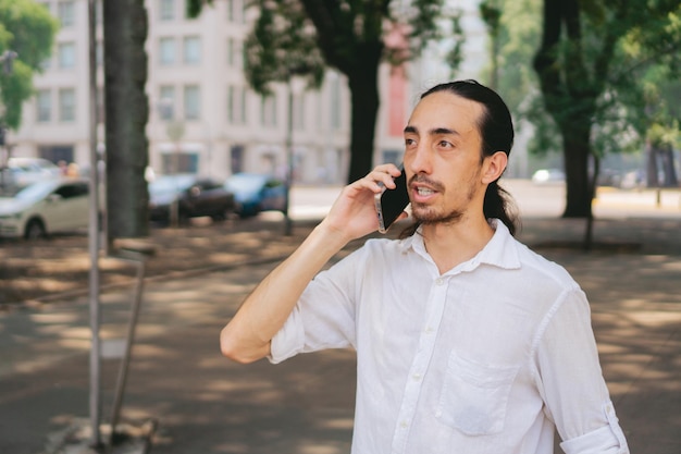 Schlanker junger lateinamerikanischer Mann mit langen Haaren, der beim Spaziergang durch die Stadt mit dem Handy telefoniert Kopierraum