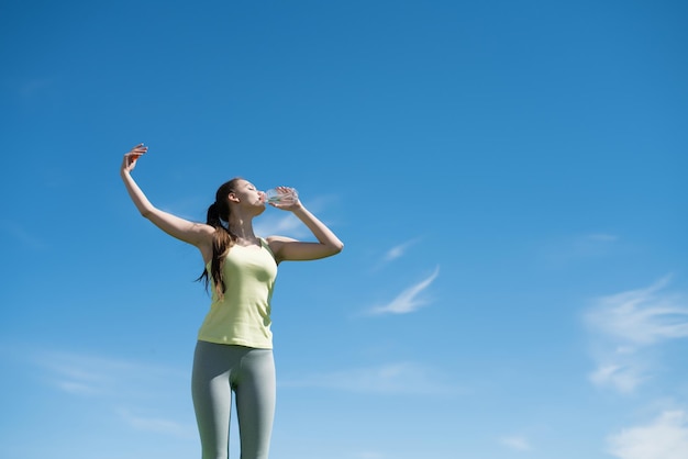 Schlankes, aktives junges Mädchen treibt Sport unter blauem Himmel, trinkt Wasser und bekommt Kraft