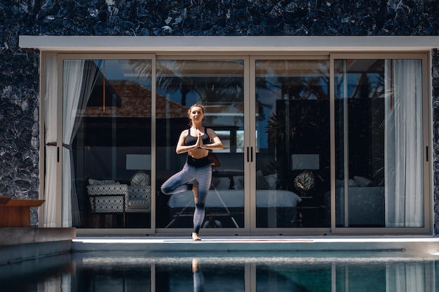 Schlankes, ruhiges Mädchen, das Sportkleidung trägt und Yoga neben dem Pool im Hintergrund ihrer Strecke macht. Sportkonzept