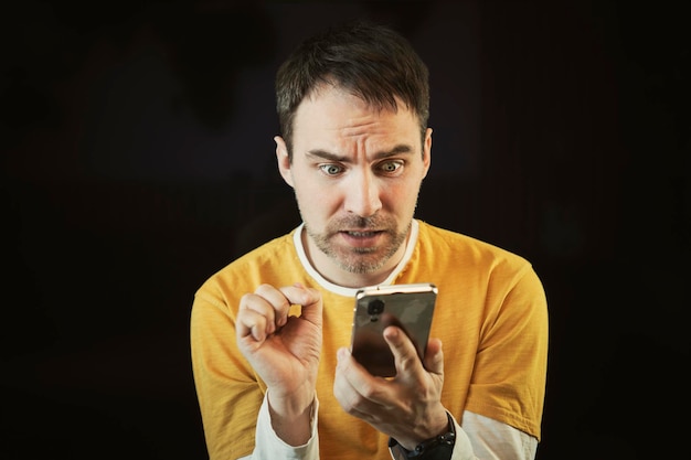 Schlechte Nachrichten im Smartphone Ein Mann schaut mit großen Augen und offenem Mund auf eine Nachricht auf seinem Handy Er ist schockiert und verängstigt Menschliche Emotionen auf schwarzem Hintergrund
